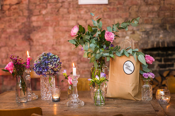 Wooded table with brown paper bag with "The Social Kitchen" label, flowers and candles. 