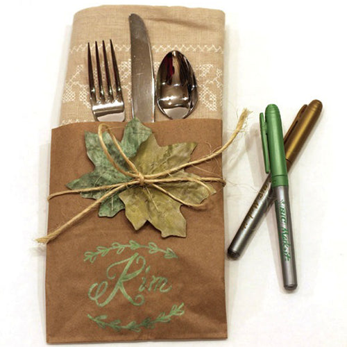 Top view of silverware wrapped in a brown napkin, tied with twine and a leaf. Designed with markers written name, "Ria" and greenery. 