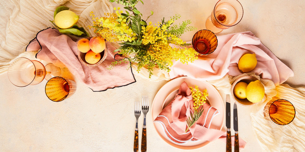 Top view of table setting and stemware with light pink napkins surrounded by lemons and flowers. 