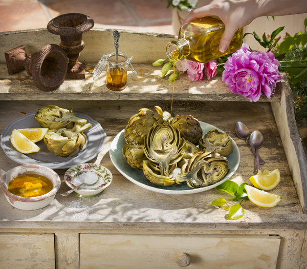 Grilled Artichokes with a person drizzling olive oil on top on wooded table top. 