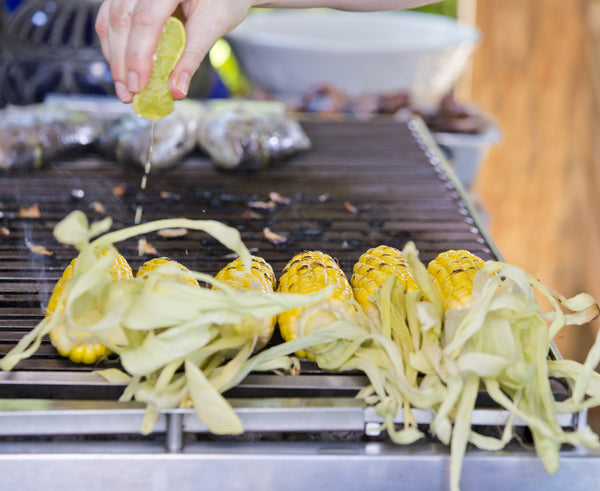 Corn being grilled on grill with hand squeezing lemon on top. 