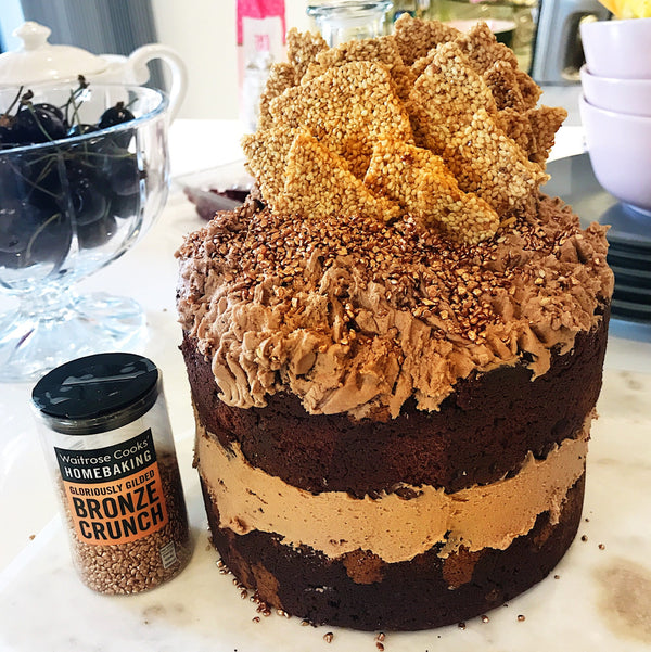 Two tiered chocolate cake with frosting and sesame snacks. On the side is a jar of Bronze Crunch. 