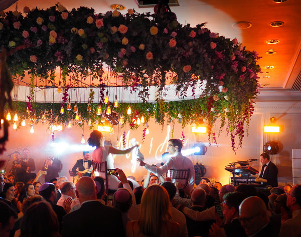 Bride and groom dancing under flower chandelier surrounded by group of friends. 