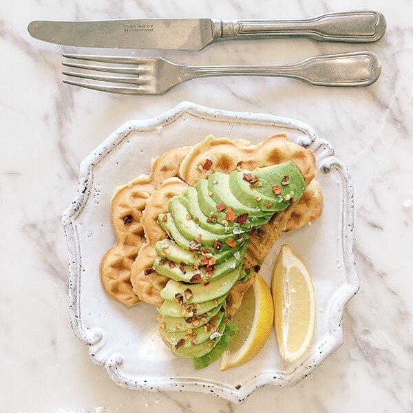 Top view of waffles topped with avocado and lemon on white dish with white marbled table in background and silverware.