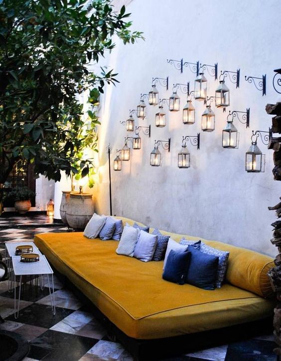 Yellow outdoor couch with mixed blue pillows surrounded by greenery and multiple lights on white wall.