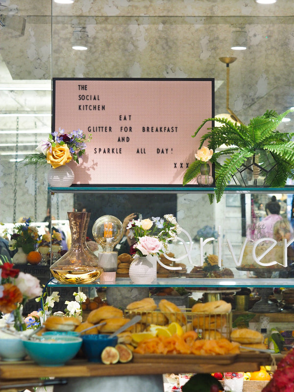 Shelves filled with baked goods and flowers. The Social Kitchen sign in background with text: Eat glitter for breakfast and sparkle all day! XXX