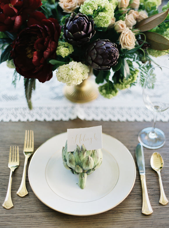 Top view of table setting on wood table with a white tablecloth with an artichoke and flowers.