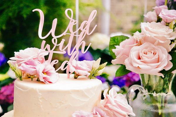 Cake topper with text, "Wed with Ted" on a white cake surrounded by flowers. 