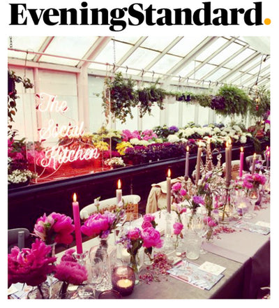 Top text: EveningStandard. View of long table filled with flowers and candles surrounded by The Social Kitchen pink text light and greenery. 