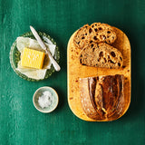 Top view of bread loaf sliced on cutting board with knife, butter and salt on green door.