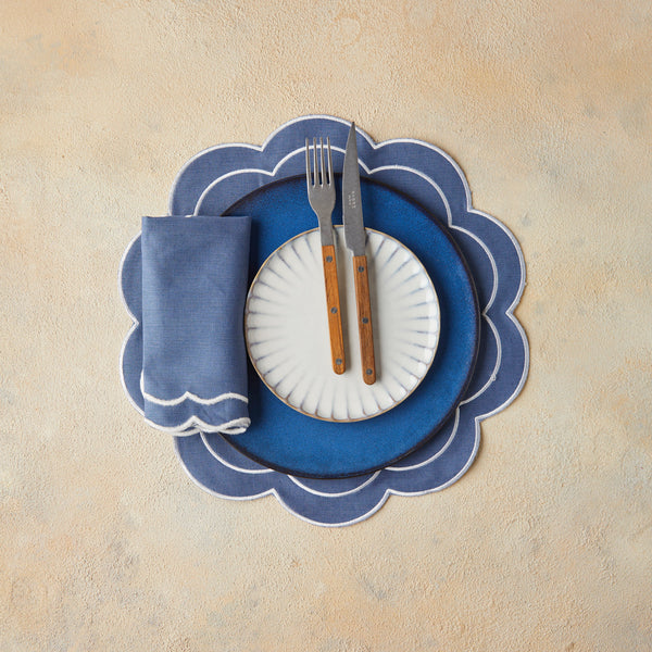 Top view of blue and white table setting with brown cutlery, blue scallop napkin, on blue scallop placemat and beige background.